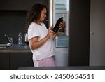 Portrait of a woman suffering from dipsomania, taking out a bottle of wine from the fridge while waking up in the morning