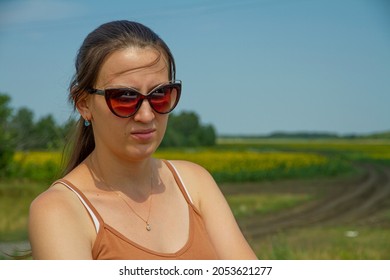 Portrait Of A Woman In Sunglasses. A Woman In Bad Mood Stands On A Sunny Summer Day In The Field. Summer Time Heat And High Temperature Concept.