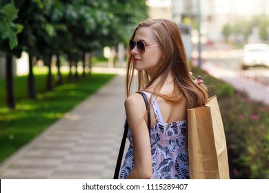 Portrait Of Woman In Sunglasses Holding Paper Bag Walk On Street. Blurry Street Background. Shopping Concept