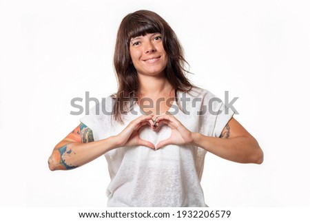 Portrait of a woman with tattoos, making a heart gesture with her hands and smiling. White background. Copy space. The concept of world heart day.