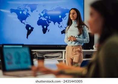 Portrait of a woman team leader presenting project in a boardroom and standing next to big screen with a world map and points on it. Business woman holding a meeting about important project. - Powered by Shutterstock