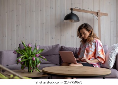 Portrait Of Woman Typing On Keyboard Of Laptop And Sitting On Soft Sofa In Casual Clothes Near Table With Vase Of Fading Tulips. Living Room In Elegant Design With Aesthetic Decor, Free Copy Space