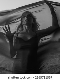 Portrait Of Woman Under Transparent Textile On Sand Dune. Girl Sneaking Through Fabric. Black And White