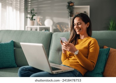 Portrait of a woman using laptop and a phone and checking email or news online while sitting on sofa at home. Searching for friends in internet social networks or working on computer. Copy space.