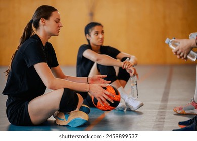 Portrait of a woman volleyball players sitting on a floor on an indoor volleyball court and having a break after the training or a match. Diverse female teammates relaxing and talking to each other. - Powered by Shutterstock