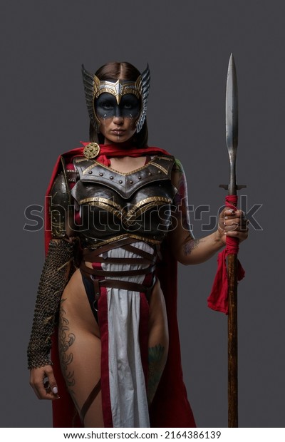Portrait of woman warrior with\
tattooed body and spear dressed in light armor staring at\
camera.