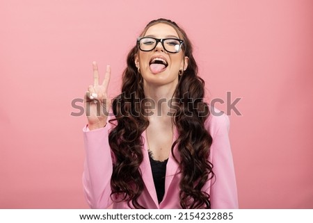 Portrait of a woman wearing glasses with a funny face sticking tongue out of mouth, teasing avoiding gaze, misbehaving, wearing a pink jacket, isolated on a pastel background.