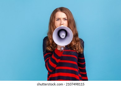 Portrait Of Woman Wearing Striped Casual Style Sweater, Taking In Megaphone, Looking At Camera With Serious Expression, Important Announcement. Indoor Studio Shot Isolated On Blue Background.