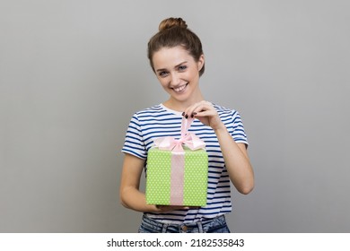 Portrait Of Woman Wearing Striped T-shirt Opening Gift Box, Unwrapping Birthday Present For Celebrating Holiday, Pulling Ribbon, Looking At Camera. Indoor Studio Shot Isolated On Gray Background.