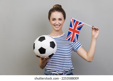 Portrait of woman wearing striped T-shirt holding british flag and black and white ball, cheering for his favorite team, supporting. Indoor studio shot isolated on gray background. - Powered by Shutterstock