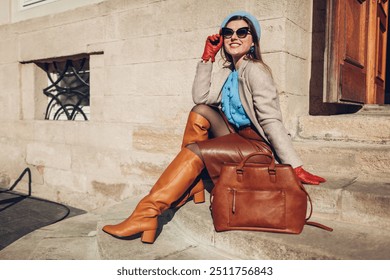 Portrait of woman wearing stylish orange boots coat beret sitting on stairs by handbag outdoors. Fall female fashionable accessories. Vintage outfit - Powered by Shutterstock