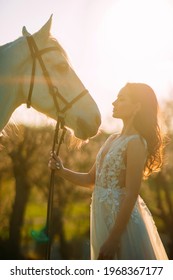 Portrait Of Woman In White Dress Near White Horse At Sunset. Backlight.