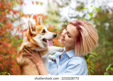 Portrait Of A Woman Who Holds Her Beloved Pembroke Welsh Corgi Dog In Her Arms, In An Autumn Park, Against A Blurred Background Of Autumn Foliage. Animal Protection, Dog Walking, Dog-human Friendship.