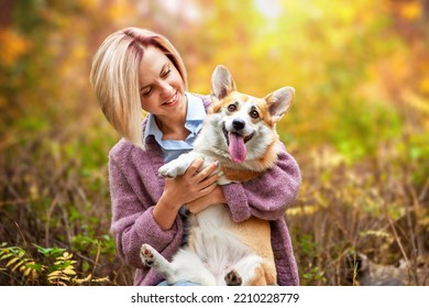 Portrait Of A Woman Who Holds Her Beloved Pembroke Welsh Corgi Dog In Her Arms, In An Autumn Park, Against A Blurred Background Of Autumn Foliage. Animal Protection, Dog Walking, Dog-human Friendship.