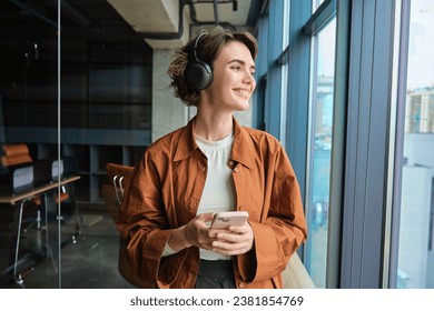 Portrait of woman working in office, digital nomad in casual clothes, wearing wireless headphones, holding mobile phone, looking outside window with pleased smile. - Powered by Shutterstock