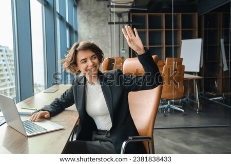 Portrait of woman working on laptop, businesswoman saying hello, waving at co-worker in her office and smiling, saying hello to colleague.