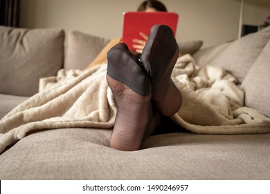 A portrait of a woman's feet in pantyhose while she is lying and resting in a couch under a warm and cosy fleece blanket reading and surfing on her tablet.
