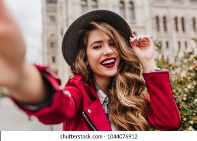 Portrait of wonderful white female model with bright makeup expressing energy in good day in Europe. Lovely curly woman in stylish hat making selfie while walking past old building.