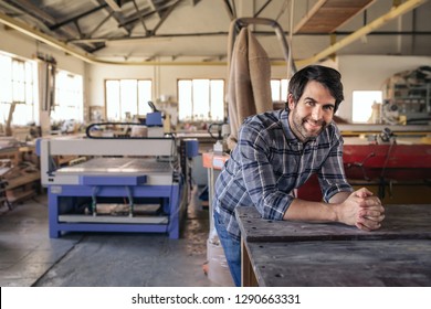 Portrait Of A Woodworker Smiling While Leaning On A Workbench In His Large Woodworking Studio Full Of Tools And Equipment 