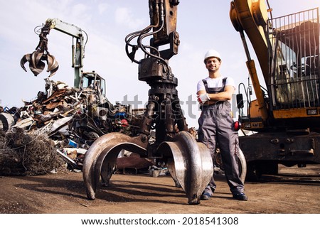 Portrait of worker standing by hydraulic industrial machine with claw attachment used for lifting scrap metal parts in junk yard.