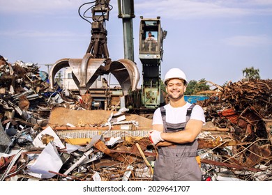 Portrait Of Worker Standing In Metal Junk Yard With Crane Lifting Scrap Metal For Recycling.