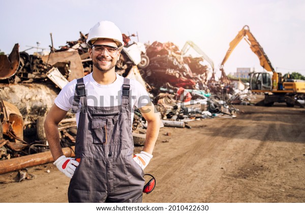 Portrait of worker standing in metal\
junk yard with crane lifting scrap metal for\
recycling.