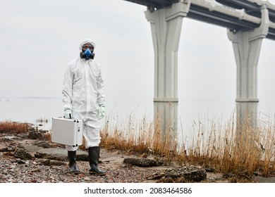 Portrait of worker wearing hazmat suit walking by water outdoors and carrying samples case, industrial waste concept, copy space
