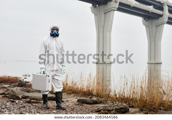 Portrait of worker wearing hazmat suit walking by\
water outdoors and carrying samples case, industrial waste concept,\
copy space