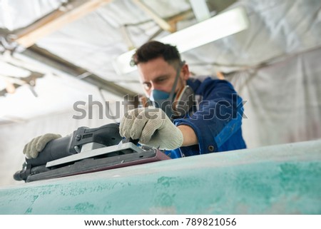Portrait of worker wearing respirator repairing boat in yacht workshop using electric polishing tool, focus on foreground