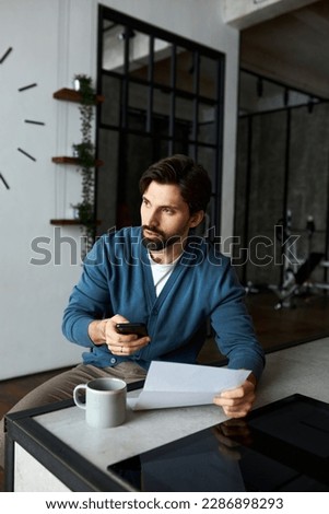 Portrait of worried concerned husband sitting at kitchen counter holding paper document, notification letter and smartphone. Handsome stylish guy dialing number after examining utility bill