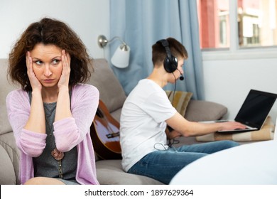 Portrait Of Worried Mother At Home On Background Of Teenage Son Carried Away With Laptop. Concept Of Youth Addiction Of Computer Games And Social Networks..