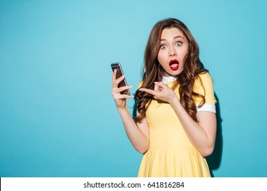 Portrait of a worried pretty girl in dress pointing at mobile phone and looking at camera isolated over blue background