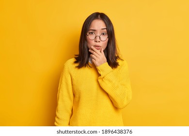 Portrait Of Worried Surprised Asian Woman Holds Chin Looks Concerned At Camera Wears Transparent Glasses And Sweater Isolated Over Yellow Background. People Face Expressions And Reaction Concept