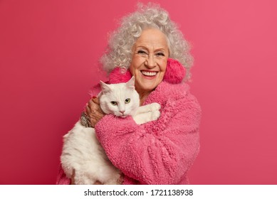 Portrait of wrinkled grandmother holds white cat on hands, stays at home during pandemic outbreak, wears fluffy earrings, soft robe, going to feed pet, isolated on pink background. Woman on pension