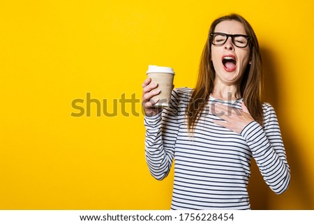 Portrait of a yawning young woman holding a paper cup with a coffee on a yellow background