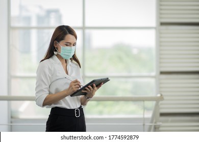 Portrait Of A Young Adult Asian Business Woman In White Shirt And Black Long Pants Wears Surgical Facemask Looking On Tablet In Company Building During VOCID-19 Pandemic. Business Stock Photo.