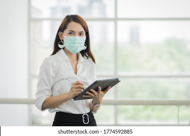 Portrait Of A Young Adult Asian Business Woman In White Shirt And Black Long Pants Wears Surgical Facemask Looking On Tablet In Company Building During VOCID-19 Pandemic. Business Stock Photo.