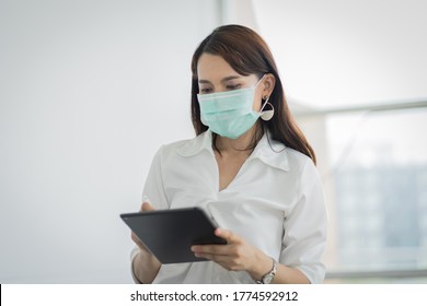 Portrait Of A Young Adult Asian Business Woman In White Shirt And Black Long Pants Wears Surgical Facemask Looking On Tablet In Company Building During VOCID-19 Pandemic. Business Stock Photo.