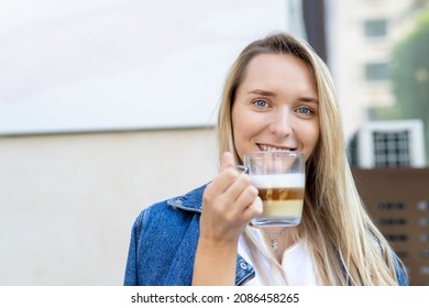 Portrait Of Young Adult Attractive Happy Woman Enjoy Smiling And Hold Cup Drinking Hot Cappuccino Coffee Against Street Cafe Wall In Morning Outdoors. Pretty Female Person Wera Jeans Jacket Outside