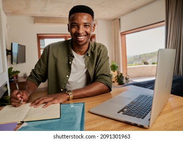 Portrait Of Young Adult Black Male Writing Note In Notebook Next To Laptop. Smiling At Camera, Sitting In Modern Apartment Working From Home.