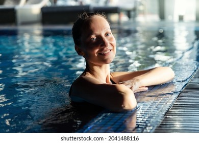 Portrait of young adult caucasian beautiful female model in sport black swimsuit in clear blue water edge indoor swimming pool at dark evening time. Relax harmony and wellness spa therapy