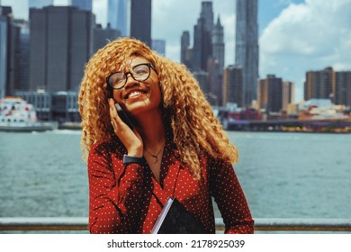 Portrait Young Adult Entrepreneur Millennial Woman With Eyeglasses And Afro Hair Smiling Talking On A Phone Call Outdoors With Manhattan New York City Skyline Behind Hudson River