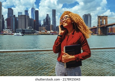 Portrait Young Adult Entrepreneur Millennial Woman With Eyeglasses And Afro Hair Talking On A Phone Call Outdoors With Manhattan New York City Skyline Behind Hudson River