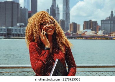 Portrait Young Adult Entrepreneur Millennial Woman With Eyeglasses And Afro Hair Talking Smiling On A Phone Call Outdoors With Manhattan New York City Skyline Behind Hudson River