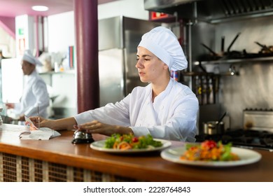 Portrait of young adult female chef cook checking orders and handing plate with meal at restaurant kitchen - Powered by Shutterstock