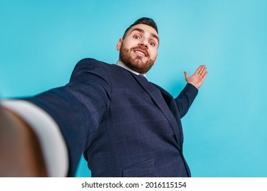 Portrait Of Young Adult Handsome Businessman In Elegant Suit Taking Selfie, Looking At Camera POV And Wave Hand, Point Of View Of Photo. Indoor Studio Shot Isolated On Blue Background.