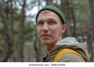 Portrait Of A Young Adult Male Walking In The Woods. Male Wearing A Green Jacket And Hat With A Yellow Backpack Behind Him. Selective Focus.