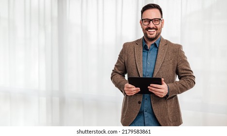 Retrato de un joven adulto sonriente, alegre empresario empresario, parado en un fondo gris, tableta de mano y mirando a la cámara.
