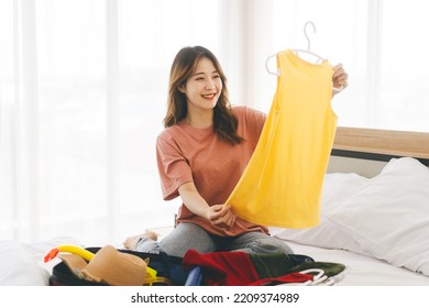 Portrait Of Young Adult Southeast Asian Dyed Hair Woman With Travel Luggage. Packing A Backpack For Journey Trip. Sitting On Bed At Home Or Hotel Bedroom.