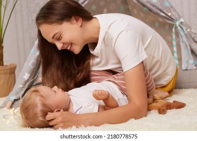 Portrait Of Young Adult Woman With Dark Long Hair Playing With Toddler Baby On Floor Near Kids Wigwam, Child Laying And Looking At Mother, Family Enjoying Time Spend Together.